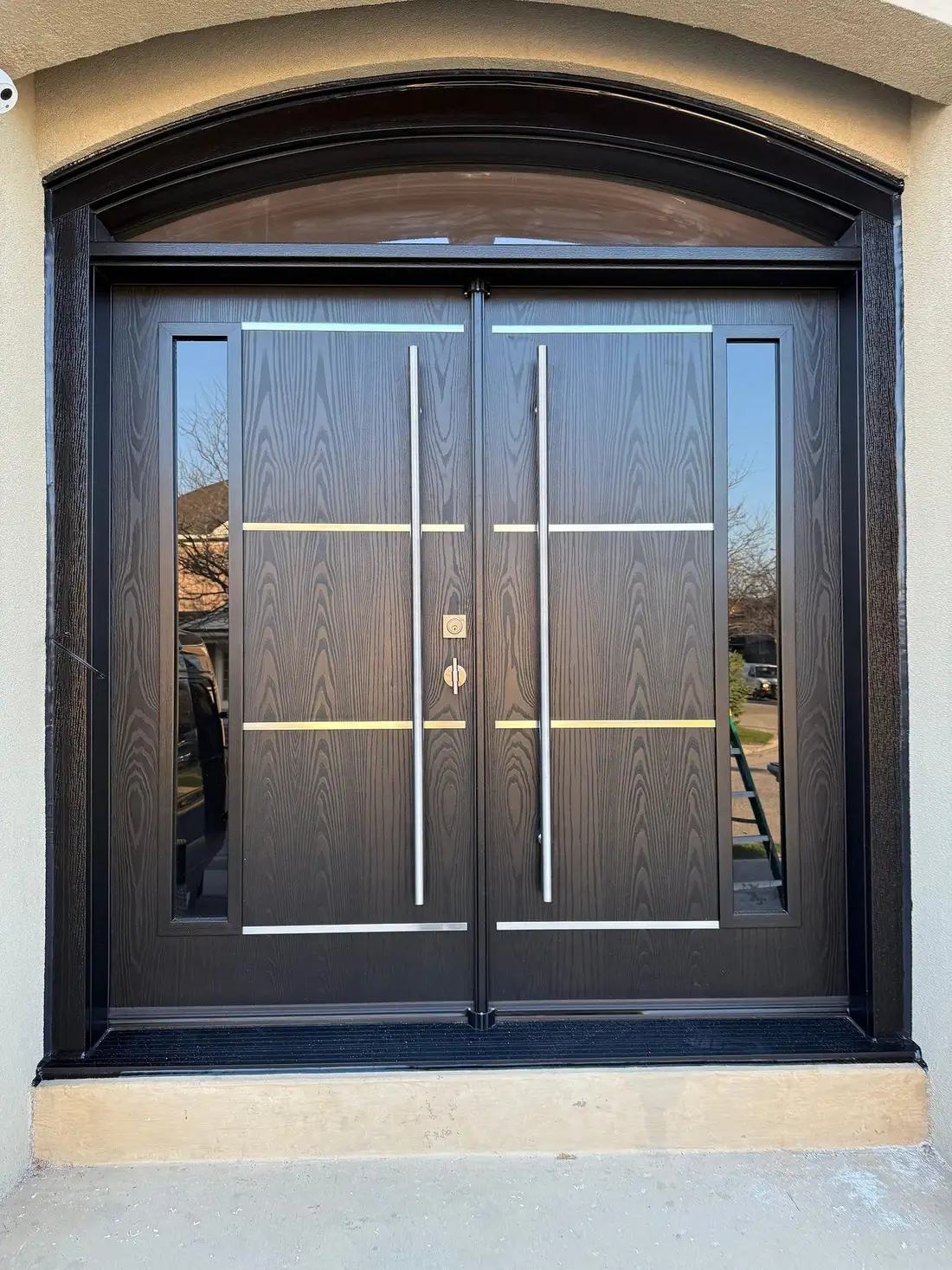 Black double entry door under an arched glass transom, with horizontal stainless bars and long pull handles, in a limestone surround