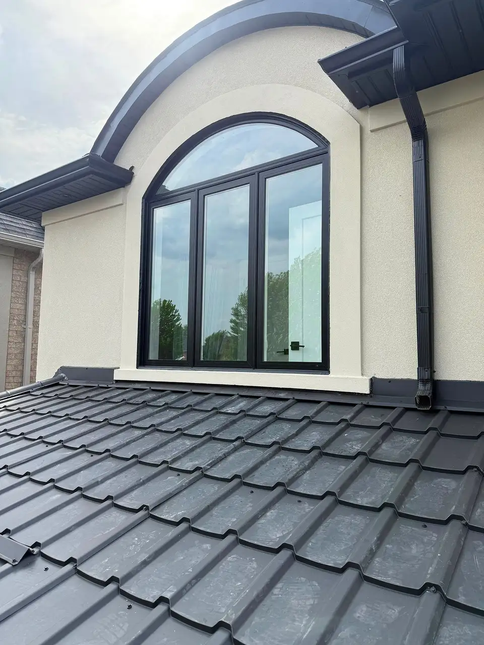 Black arched window with casement sashes, set in stucco above a metal-tile roof