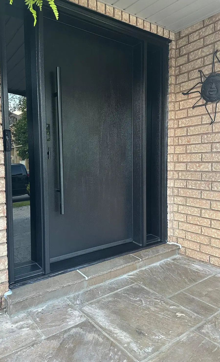 Wide black flush entry door with a full-height pull and a glass sidelite, in a buff brick porch