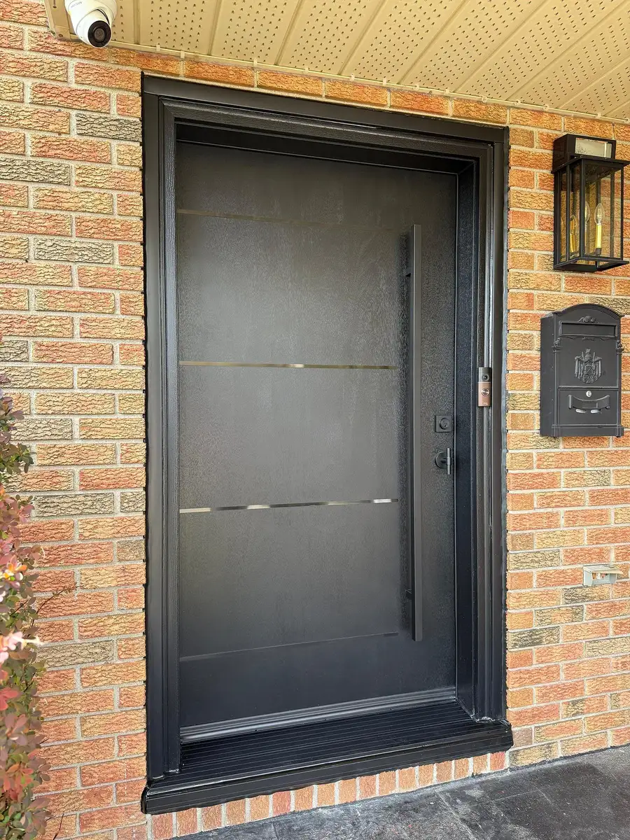 Black flush entry door with thin horizontal metal inlays and a vertical pull, in a brick entrance with black wall lanterns