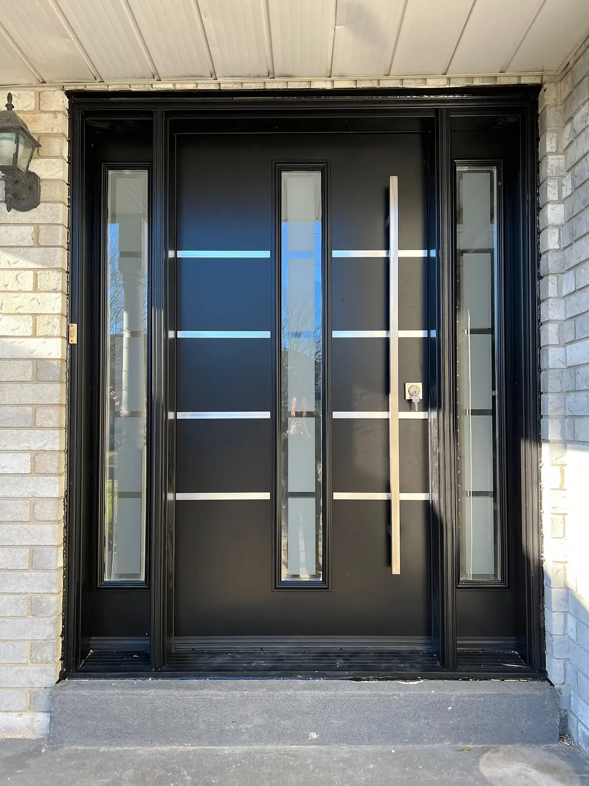Black entry door with two glass sidelites, horizontal stainless bars and a long pull, in a buff brick entrance