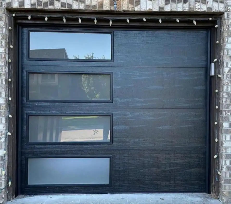 Wide black woodgrain garage door with glass lites, under a row of soffit lights