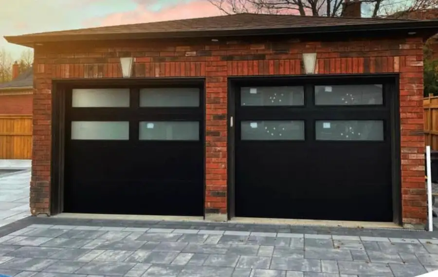 Pair of black garage doors with rows of glass lites, set in red brick above an interlock driveway