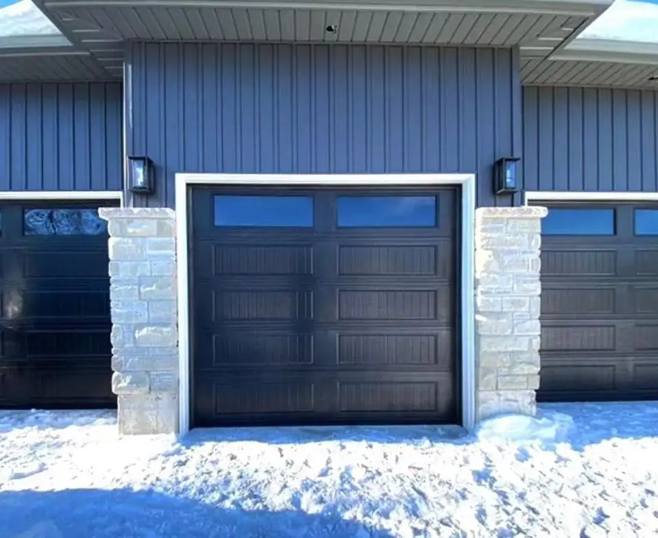 Black raised-panel garage door with a row of lites, between stone pillars on a blue house, with snow on the driveway