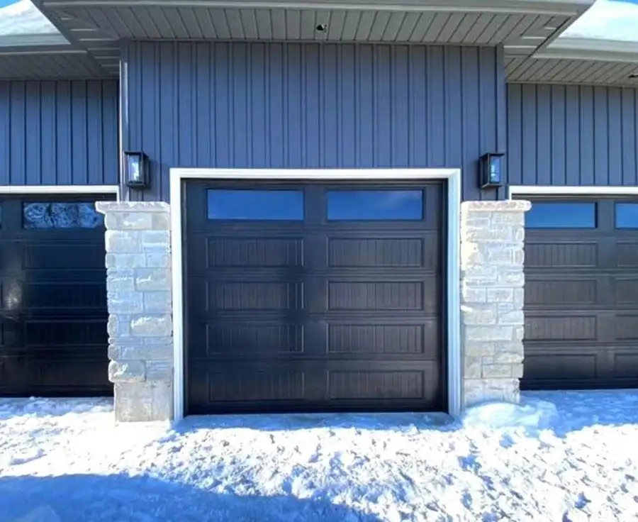 Black raised-panel garage door with a row of lites, between stone pillars on a blue house, with snow on the driveway