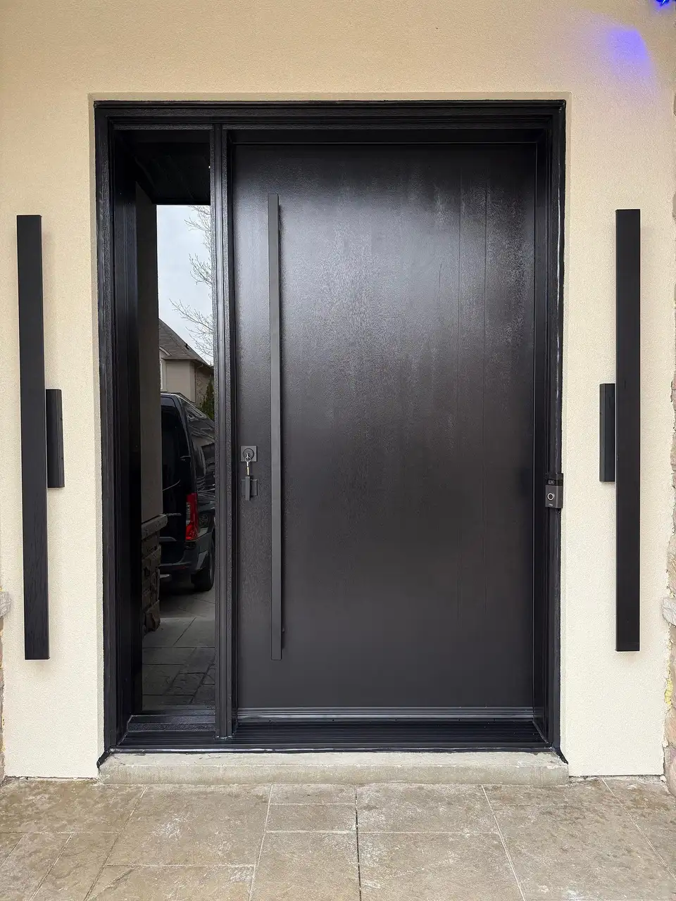 Dark fibreglass entry door with a tall square pull bar and a tinted glass sidelite, between two tall black wall lights