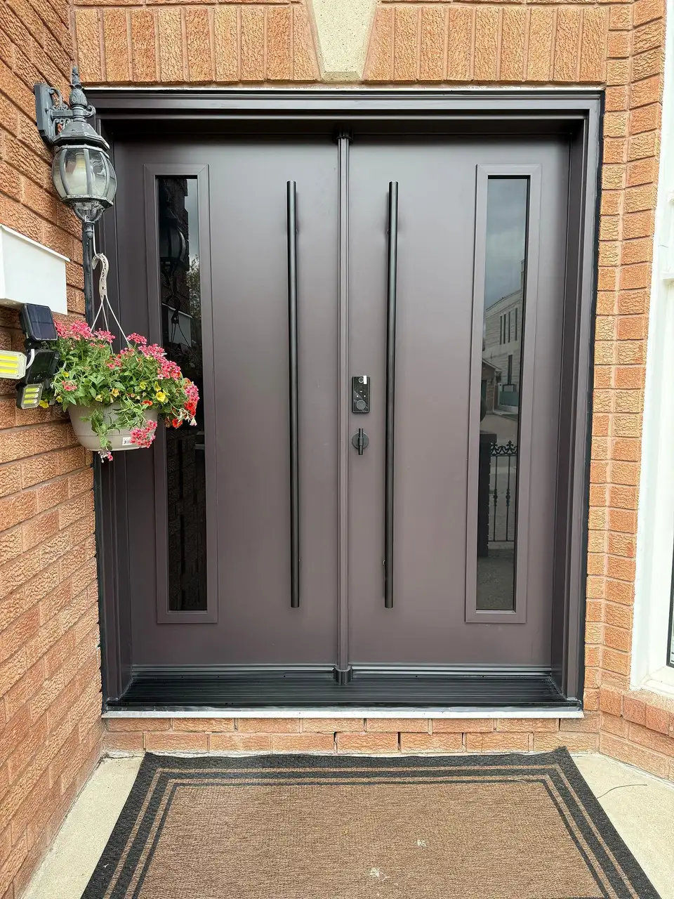 Dark double entry door with dual pull bars and slim vertical glass inserts, in red brick with a hanging flower basket