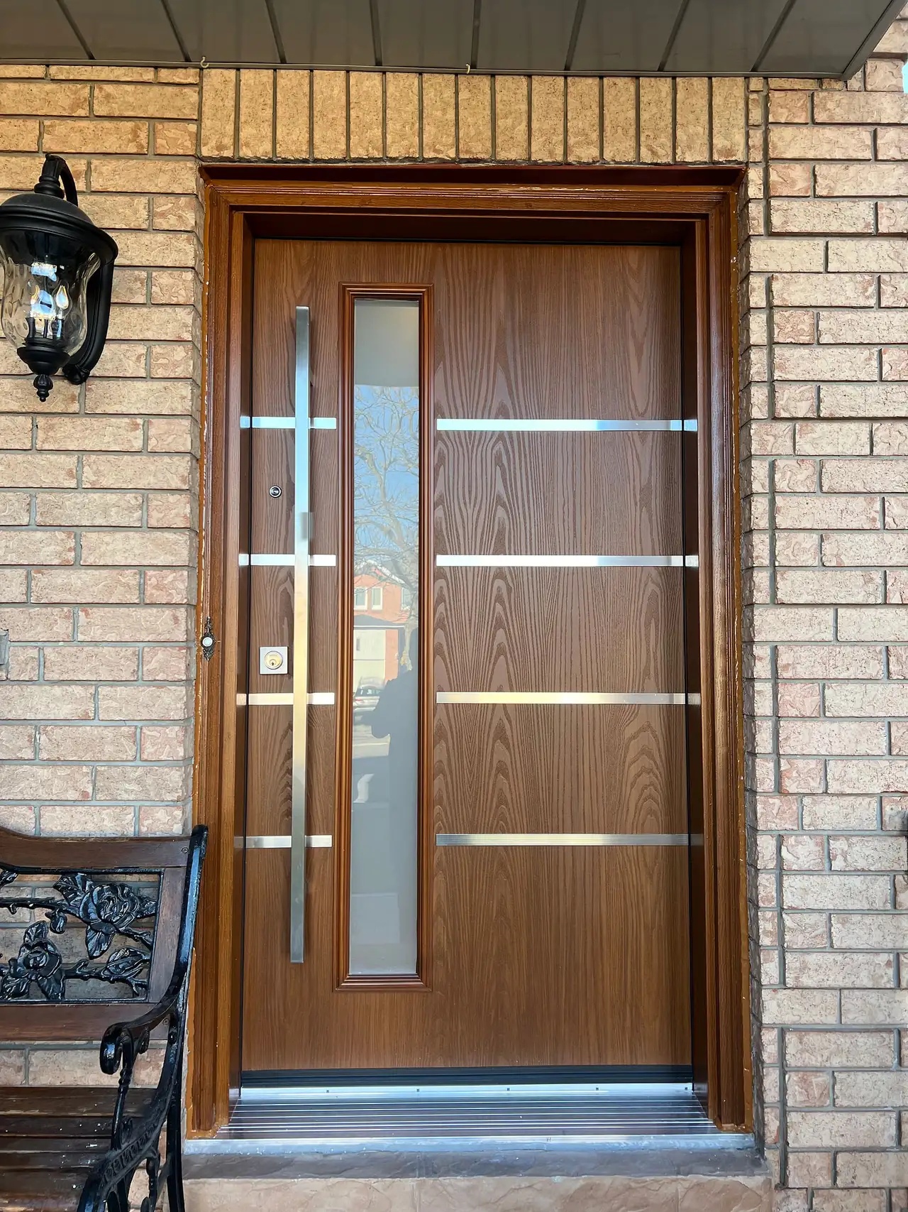 Oak woodgrain entry door with horizontal stainless bars and a glass sidelite, in a brick porch with a wrought-iron bench