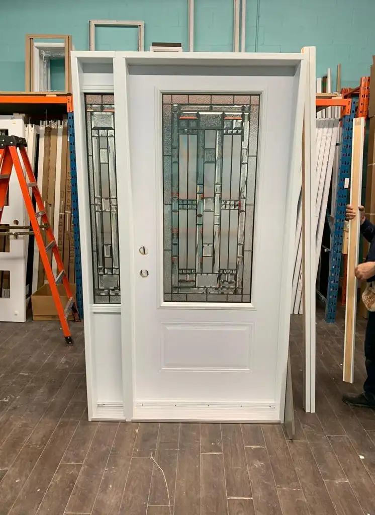 White prehung entry door with a decorative glass insert and matching sidelite, standing in the shop between racks of door slabs and frames
